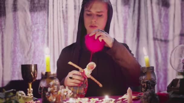 Dark magic voodoo witch performing ritual and casting spell piercing doll with needles sitting in dark room with burning candles. — Stock Video