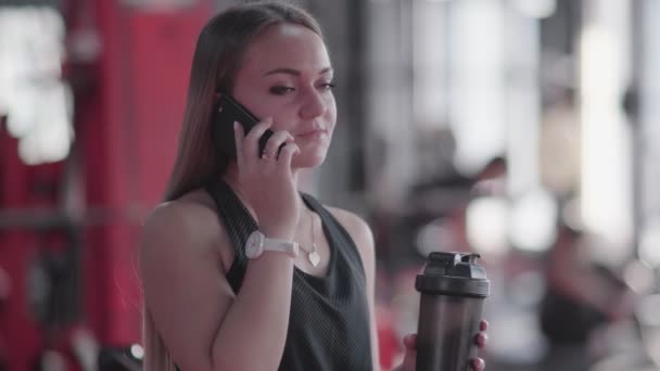 Junge blonde Frau telefoniert und hält Shaker mit Proteincocktail in der Hand — Stockvideo