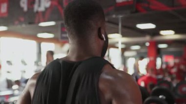 Kablosuz kulaklıklı Afrikalı Amerikalı vücut geliştirici spor salonundaki halterlerle yükseltme egzersizi yapıyor.