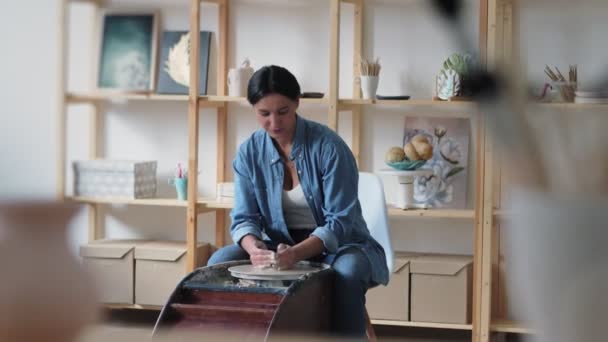 Mavi gömlekli kadın sandalyede oturuyor ve geniş profesyonel çömlekçilikte çalışırken tekerlek üzerinde kil kaplar yapıyor. — Stok video