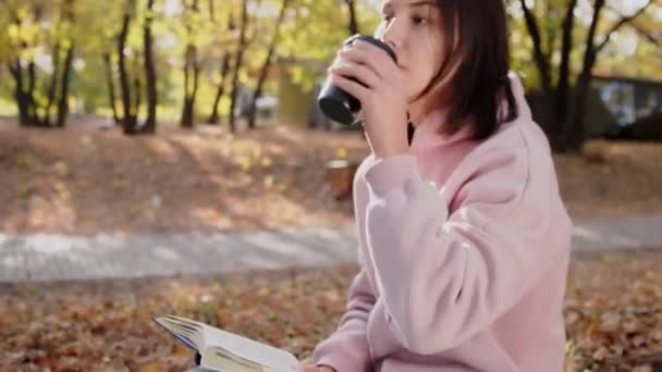 Romantic brunette woman drinks coffee and reads a book in the park at fall on a sunny day — Stock Video