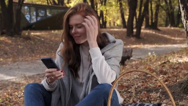 Hermosa mujer joven se sienta en la manta en el parque y utiliza el teléfono en otoño en un día soleado — Vídeo de stock