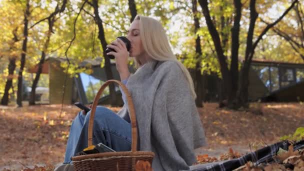 Mujer rubia joven bebe café y usa el teléfono en el parque en otoño en un día soleado — Vídeo de stock