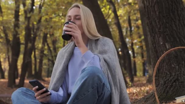 Blonde woman drinks coffee and uses phone in the park at fall on a sunny day — Stock Video