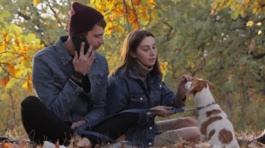 Güz parkında Jack Russell Terrier köpeği olan genç bir çift. Kadın köpekle oynarken erkek telefonda konuşur.