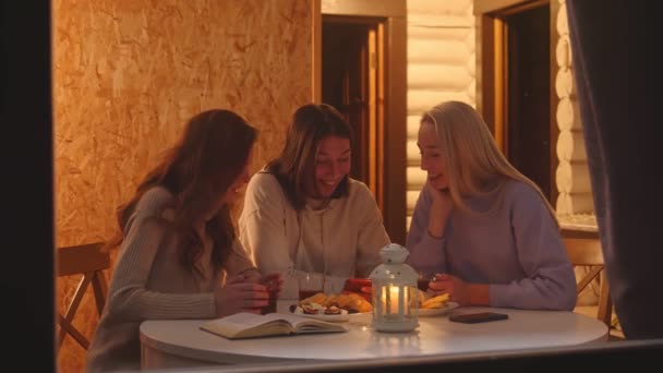 Três mulheres bebem café ou chá e olham para o telefone em casa de campo de férias de madeira à noite — Vídeo de Stock