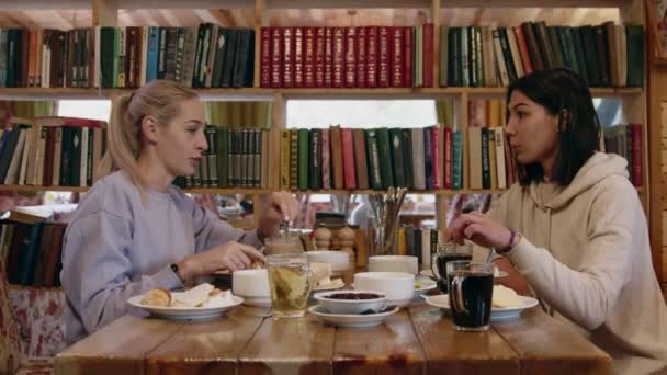 Dos mujeres jóvenes se sienta en la cafetería con estantería en el fondo — Vídeos de Stock