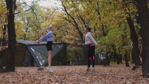 Két testhezálló szőke és barna nő tornázik az őszi parkban. — Stock videók