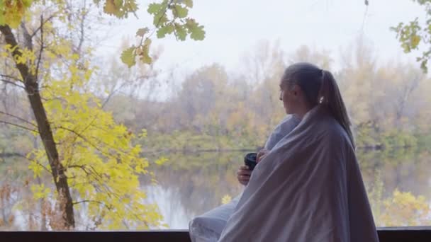 Молодая блондинка с одеялом на плечах пьет горячий чай и сидит на террасе у реки — стоковое видео
