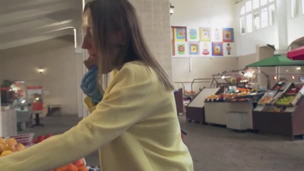 Женщина в желтом пальто, защитной маске и синих перчатках нюхает и выбирает свежие фрукты на полках магазинов на рынке — стоковое видео