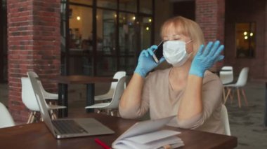 Koruyucu maskeli ve eldivenli kadın kafede defterle otururken telefonla konuşuyor.