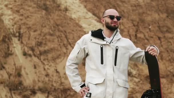 Handsome bald bearded man with sunglasses posing with sandboard in desert or sand quarry — Stock Video