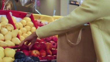 Sarı ceketli bir kadının arka görüntüsü kırmızı elmaları markette bir kese kağıdına koyar.