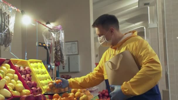 Un uomo con maschera protettiva e guanti sceglie un mandarino fresco, li mette in un sacchetto di carta ang lo dà al venditore nel mercato — Video Stock