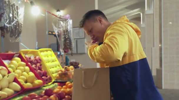 Молодой человек говорит по телефону и выбирает свежие яблоки и кладет их в бумажный пакет на рынке — стоковое видео