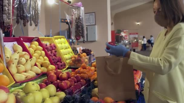 Femme en manteau jaune, masque de protection et gants bleus choisit une pomme fraîche au marché — Video