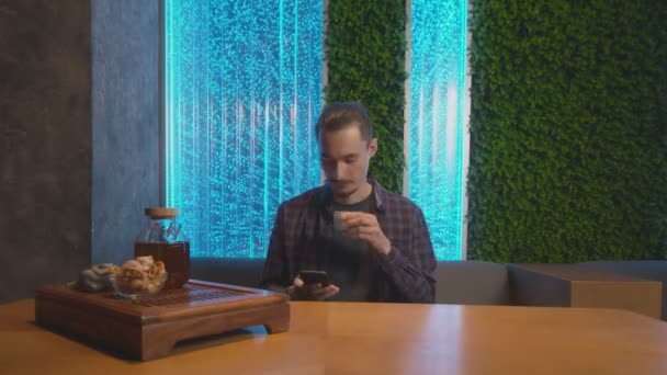 Vista frontal de homem bonito bebe chá chinês tradicional e usa telefone no café — Vídeo de Stock