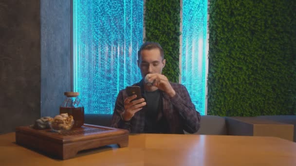 Z przodu przystojny mężczyzna pije tradycyjną chińską herbatę, je ciasteczka i używa telefonu w kawiarni — Wideo stockowe