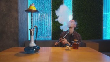 Genç hippi bir kulüpte telefon edip nargile tüttürüyor, sis bulutu üfleyerek rahatlıyor.