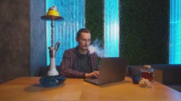 Vonzó vidám ember dolgozik notebook, miközben dohányzik egy shisha — Stock videók
