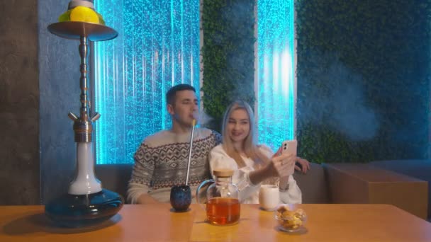 Mladý pár sedí v objetí v baru Shisha Lounge Hookah. Mladá žena fotí na svém chytrém telefonu — Stock video