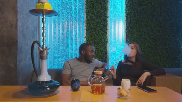 Mladý africký muž pije čaj a mluví s běloškou, zatímco ona kouří hookah — Stock video