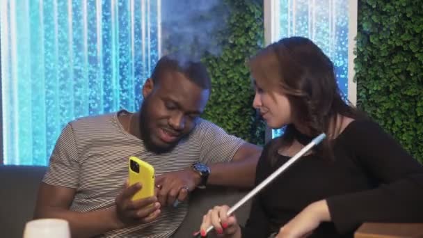 Νεαρός Αφρικανός δείχνει στην κοπέλα του κάτι μεγάλο στο κινητό του ενώ καπνίζει ναργιλέ. — Αρχείο Βίντεο