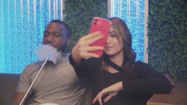 Jonge vrouw neemt selfie op telefoon, terwijl haar afrikaanse vriendje rookt hookah — Stockvideo