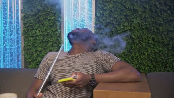 Jeune homme africain seul assis dans un sac de shisha, fume et utilise le téléphone — Video