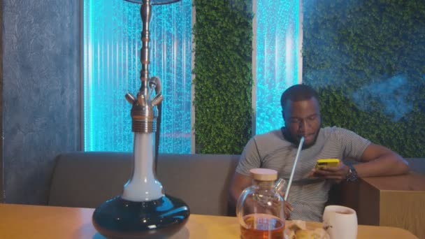 Jonge Afrikaanse man alleen zit in shisha tas, rookt en gebruikt telefoon — Stockvideo