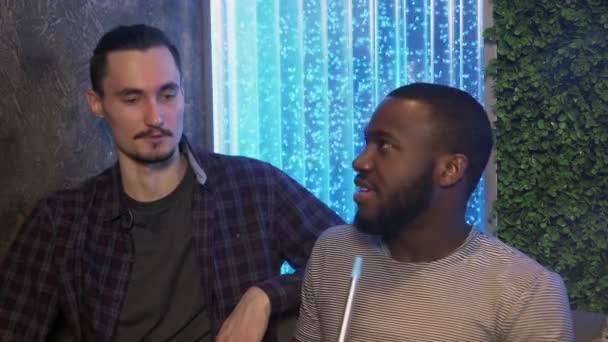 Africano se sienta en shisha bar, fuma y habla con su amigo — Vídeos de Stock