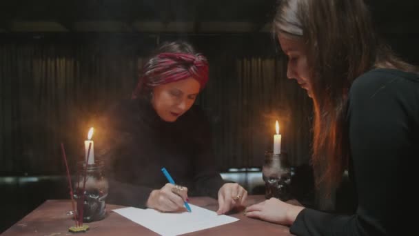 シニア女性占い師は魔法を伝える数学占いを行うためにクライアントの答えを書きます — ストック動画