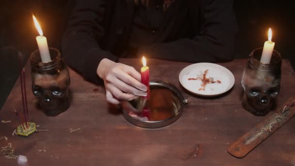 Крупным планом молодая ведьма-гадалка смотрит в зеркало на столе и держит свечу, которая капает воск в тарелку в ужасном магическом ритуале — стоковое видео