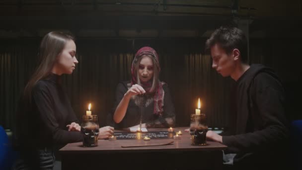 Μάγισσα γυναίκα μάντισσα προσπαθεί να μιλήσει με φάντασμα. Χρησιμοποιεί μπλε κρύσταλλο και πίνακα με γράμματα. Δύο γυναίκες και ένας άντρας χρησιμοποιούν Ouija Board Spirit Game — Αρχείο Βίντεο