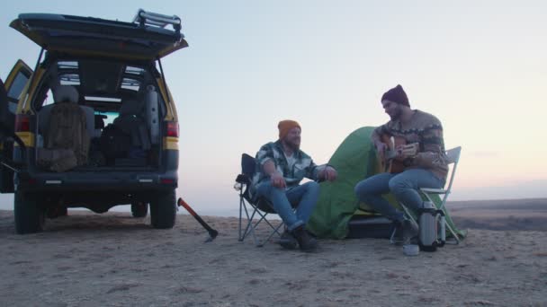 Dois amigos bonitos campistas senta-se em cadeiras campistas perto da tenda, toca guitarra e canta — Vídeo de Stock