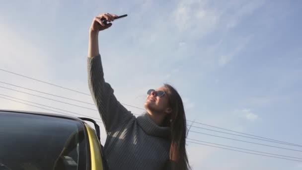 Młoda kobieta w okularach przeciwsłonecznych wychyliła się przez okno samochodu i nagrywa własne filmy z selfie — Wideo stockowe