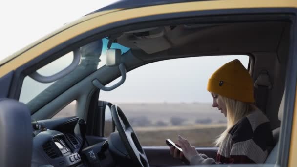 Νεαρή γυναίκα με κίτρινο καπέλο κάθεται στο αυτοκίνητο και χρησιμοποιεί το τηλέφωνο — Αρχείο Βίντεο