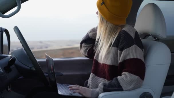 Joven freelancer en sombrero amarillo se sienta en coche y trabaja con cuaderno al aire libre — Vídeos de Stock