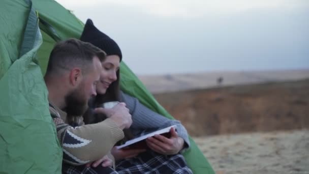 Vue latérale de yong homme et femme assis près de la tente et la voiture, boit un thé chaud et lire le livre dans la montagne — Video