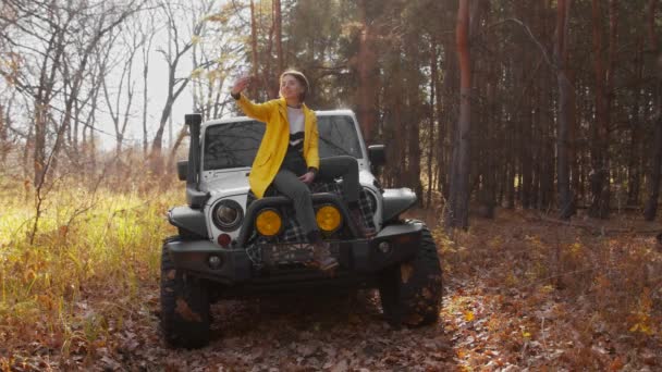 Ung glad kvinna i gul jacka sitter på bilen och tar selfie på telefon i höstskogen — Stockvideo