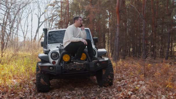 Jeune homme en pull blanc assis sur sa voiture blanche et parle au téléphone dans la forêt d'automne — Video