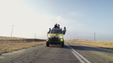 Genç çift, tatil arabasının çatısında oturuyor ve güneş gözlüklü genç kadın, sürücü arabasından sarkıyor.