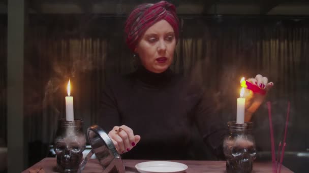 Senior bruxa cartomante mulher detém espelho em uma mão e acende vela vermelha e pingando cera em uma placa em terrível ritual mágico — Vídeo de Stock