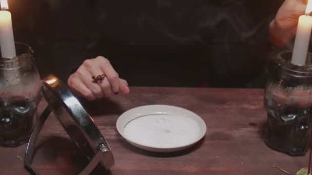Senior heks waarzegster houdt spiegel in een hand en steekt rode kaars aan en druipt was in een bord in verschrikkelijke magische rituelen — Stockvideo