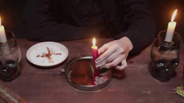 Acercamiento de la joven bruja adivina mira el espejo en la mesa y sostiene la vela que gotea cera en un plato en terrible ritual mágico — Vídeo de stock