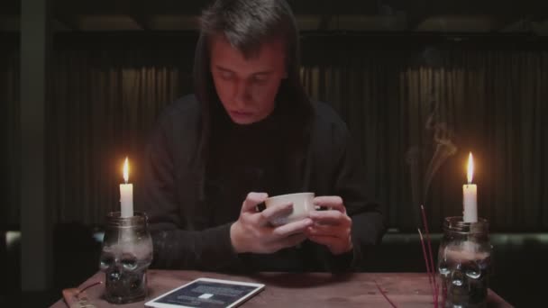 Concentrado misterioso feiticeiro cartomante masculino está segurando xícara de café e olha para tablet para ler o destino — Vídeo de Stock