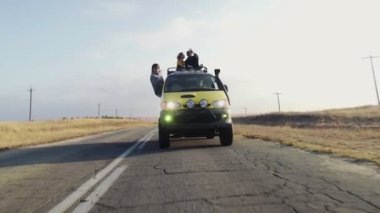 Genç çift, tatil arabasının çatısında oturuyor ve güneş gözlüklü genç kadın, sürücü arabasından sarkıyor.