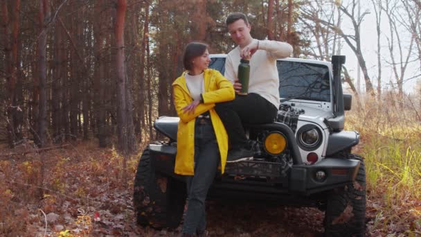 Jovem casal encantador com um carro branco na floresta de outono. Eles bebem chá da garrafa térmica — Vídeo de Stock