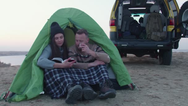 Jeune homme et jeune femme assis près de la tente et de la voiture, boit un thé chaud et lit le livre dans la montagne — Video