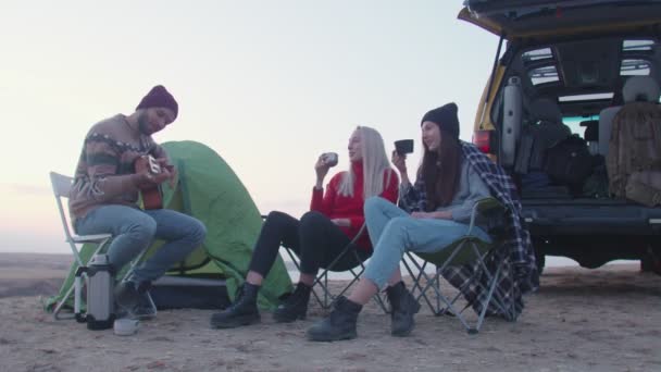 年轻人弹吉他，两个女人坐在露营椅上喝热茶 — 图库视频影像